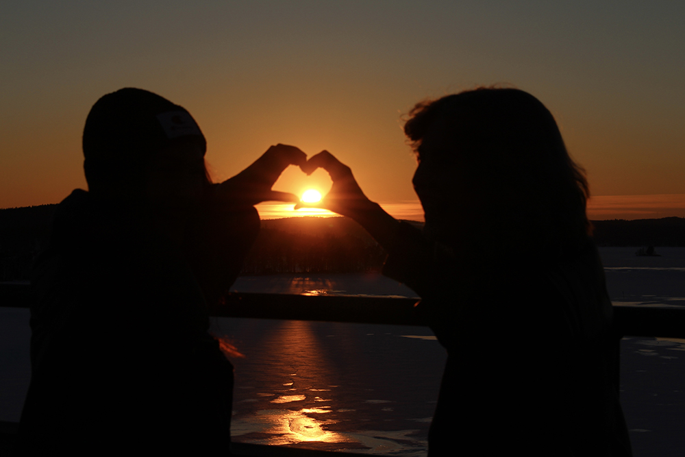 Kaksi opiskelijaa muodostavat käsillä sydämen Kärkisten sillalla auringon laskiessa.