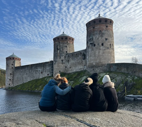 Viisi opiskelukaveria istuu kalliolla vierekkäin selät kameraan päin ja heidän takana näkyy Olavin linna