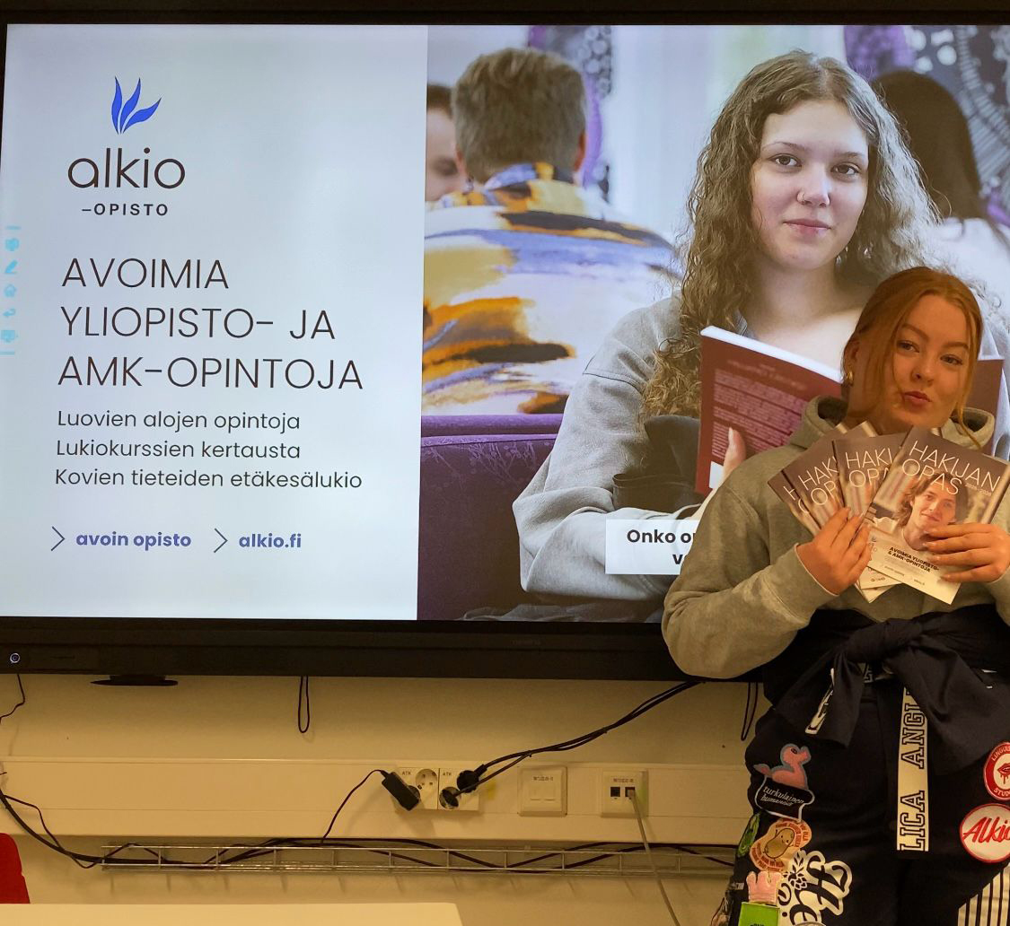 Vilma esittelemässä lukiossa Alkio-opistoa. Vilmalla on päällä yliopiston haalarit. Taustalla näkyy Alkio-opiston esittelyn ensimmäinen dia.