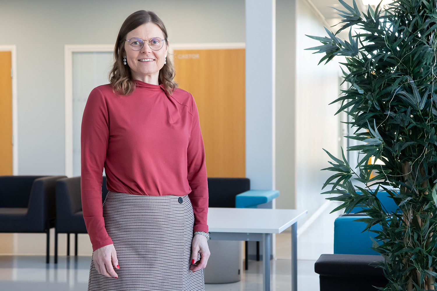 Jaana Laitila Helmi-rakennuksen Linnunrata-tilassa. Taustalla näkyy kalusteita ja oikealla puolella viherkasvi.